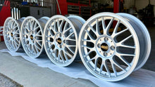 Load image into Gallery viewer, Four 16-inch BBS RG-F aluminum wheels with silver finish and black/gold logos, perfect for E30 BMWs, displayed in a garage.
